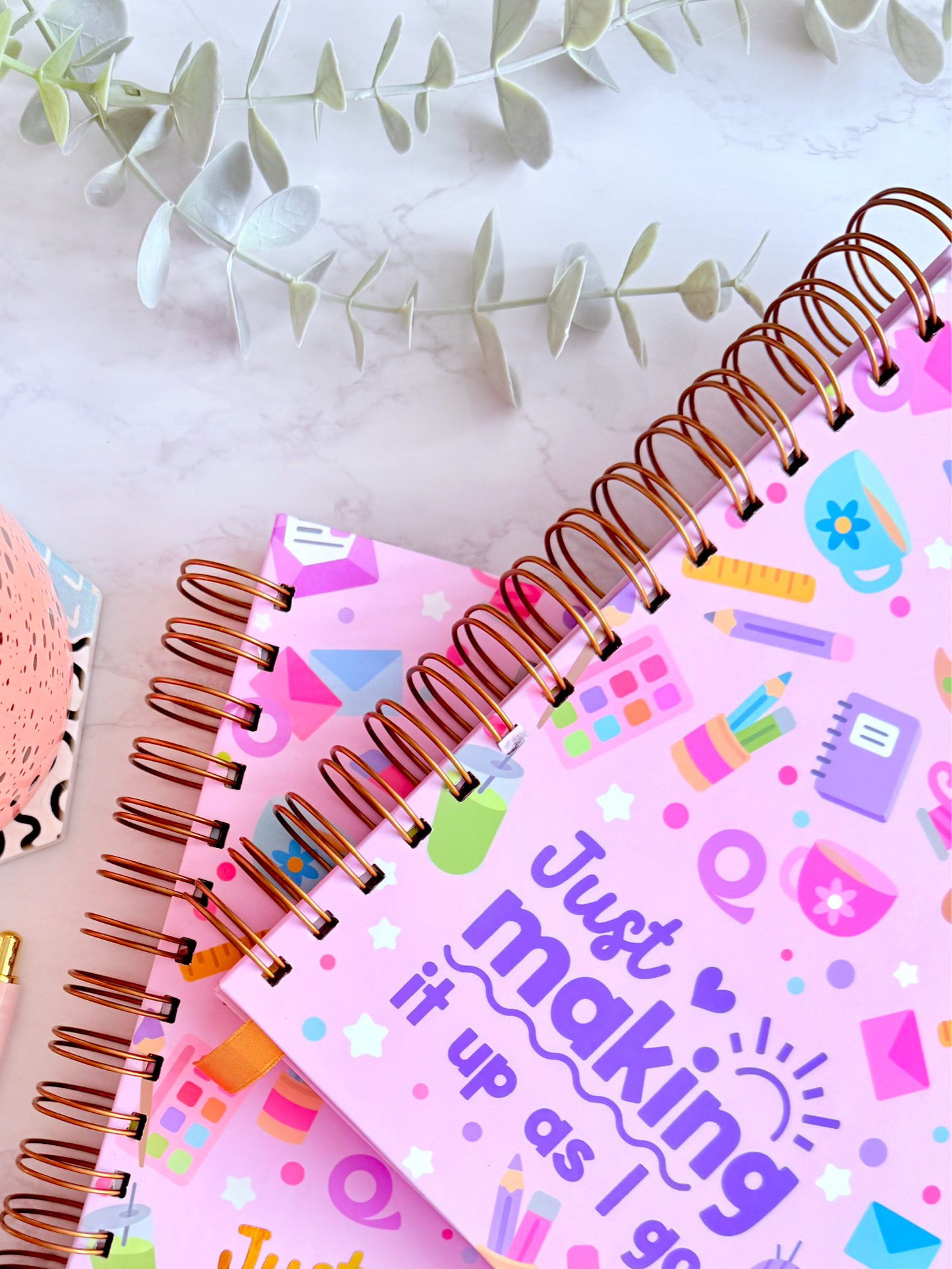 Two spiral-bound notebooks with colorful designs and text, a cup of tea, pen, and tape on a marble surface.
