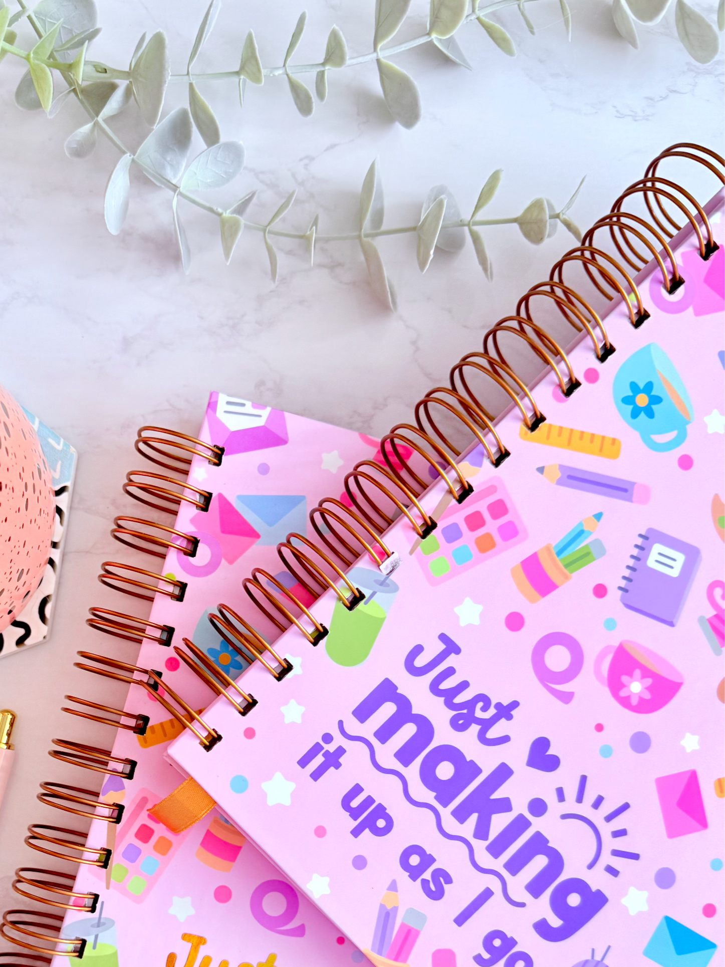 Two spiral-bound notebooks with colorful designs and text, a cup of tea, pen, and tape on a marble surface.