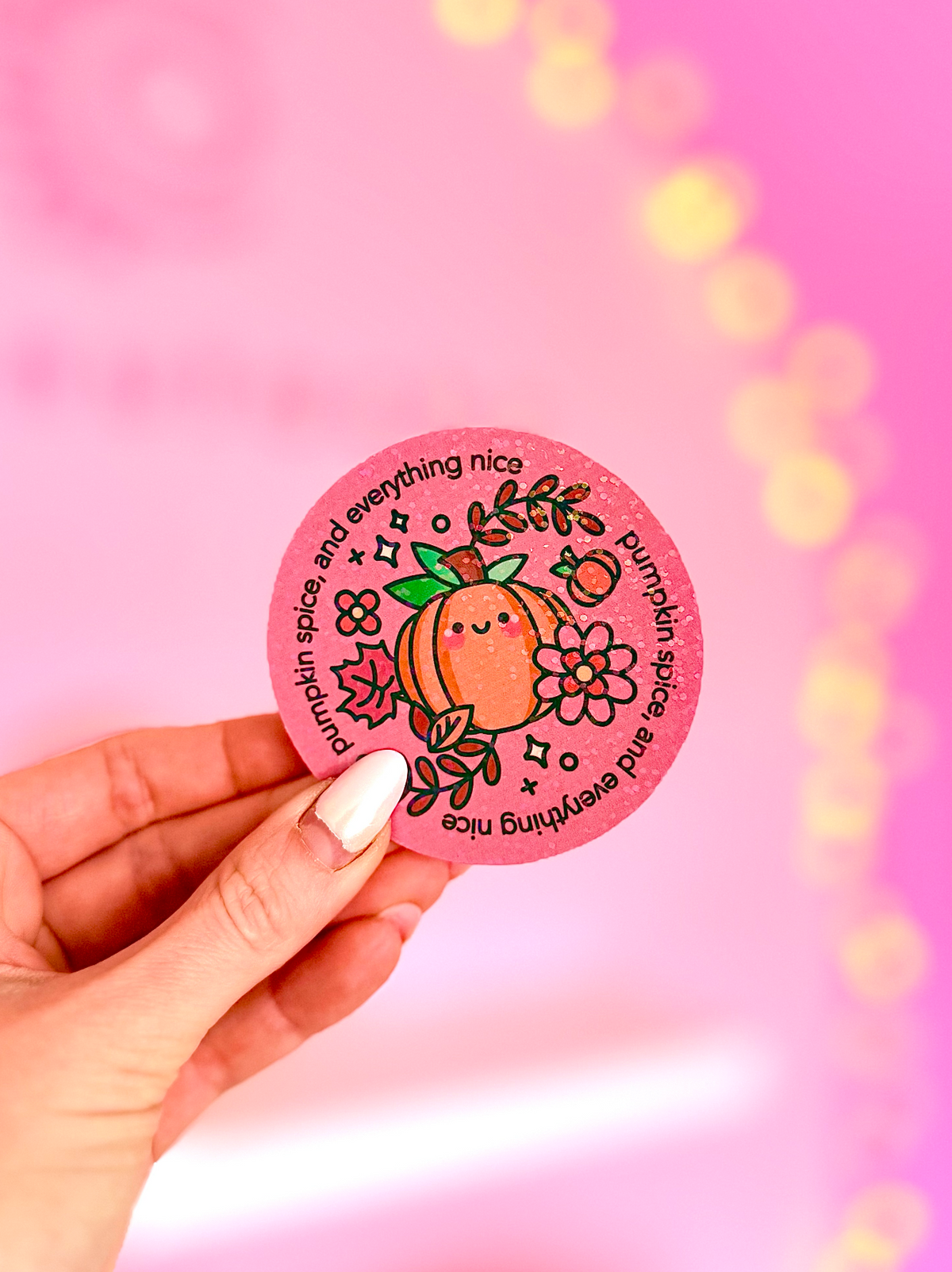 A hand with white manicured nails holds a round pink sticker featuring a cute orange character, flowers, and the text 'summer spice and everything nice' against a bright pink bokeh background.