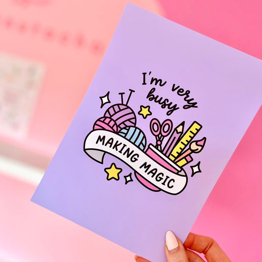 Art print with the text 'I'm BUSY MAKING MAGIC' surrounded by illustrations of scissors, yarn, and crafting tools on a pink background, held in a hand against a pink background.