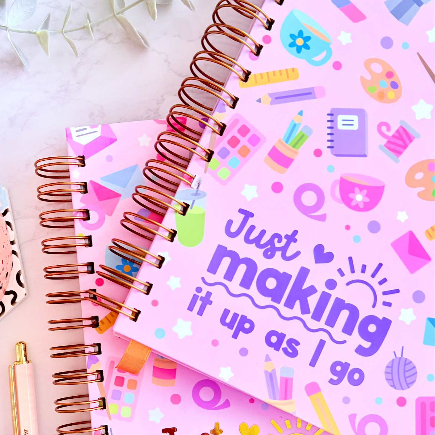 Two pink spiral-bound notebooks with colorful text and designs, a pen, and a cup on a light surface.