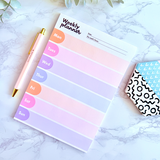 Weekly planner pad with colorful days of the week labels on a marble surface with a pen and decorative elements.