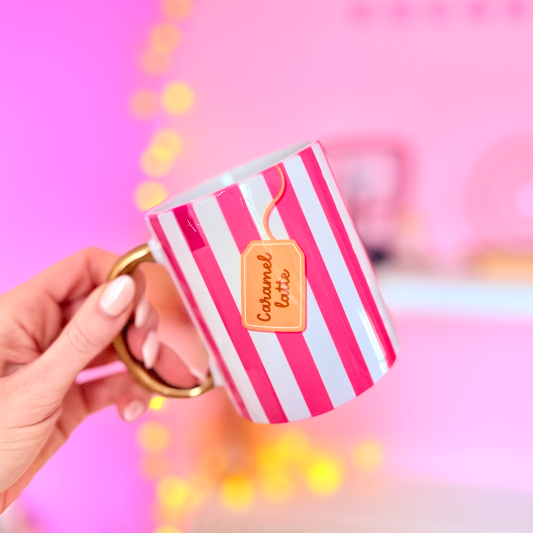 Hand holding a striped mug with 'Caramel Latte' text against a pink background