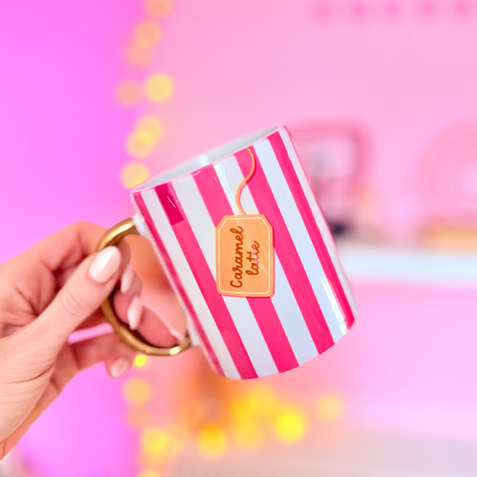 Hand holding a striped mug with 'Caramel Latte' text against a pink background