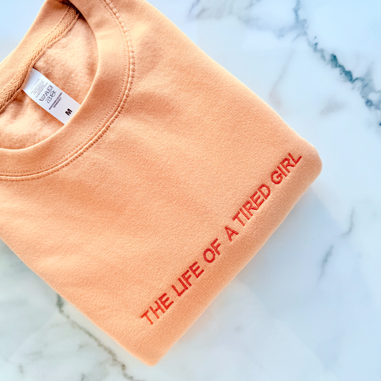 Folded peach-colored sweatshirt with 'The Life of a Tired Girl' text on a marble surface