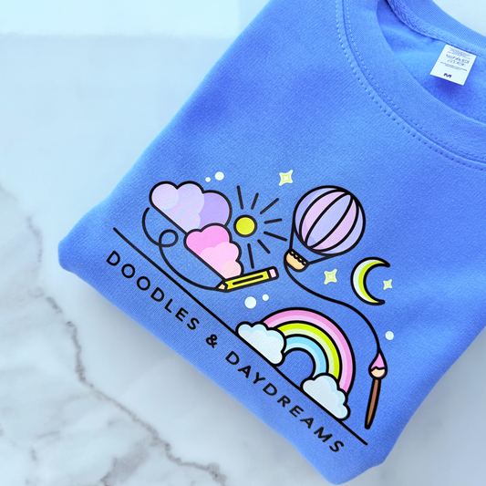 Blue sweatshirt with colorful doodles and 'Doodles & Daydreams' text on a marble surface