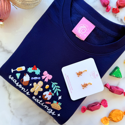 Navy blue sweatshirt with a festive embroidered design saying 'season's eatings' on a marble surface with candy accents for christmas