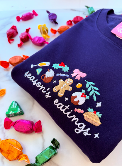 Navy blue sweatshirt with 'season's eatings' design surrounded by colorful candies on a white background