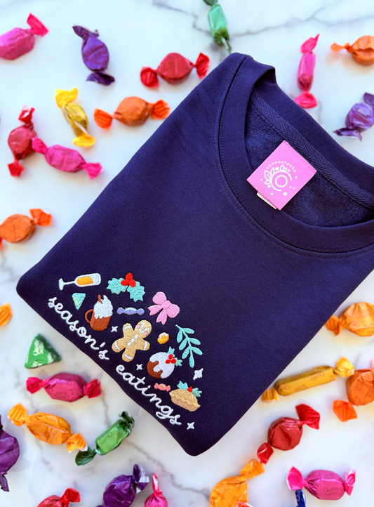 Navy blue sweatshirt with colorful graphics and text 'season's eatings' on a white background with candy decorations.