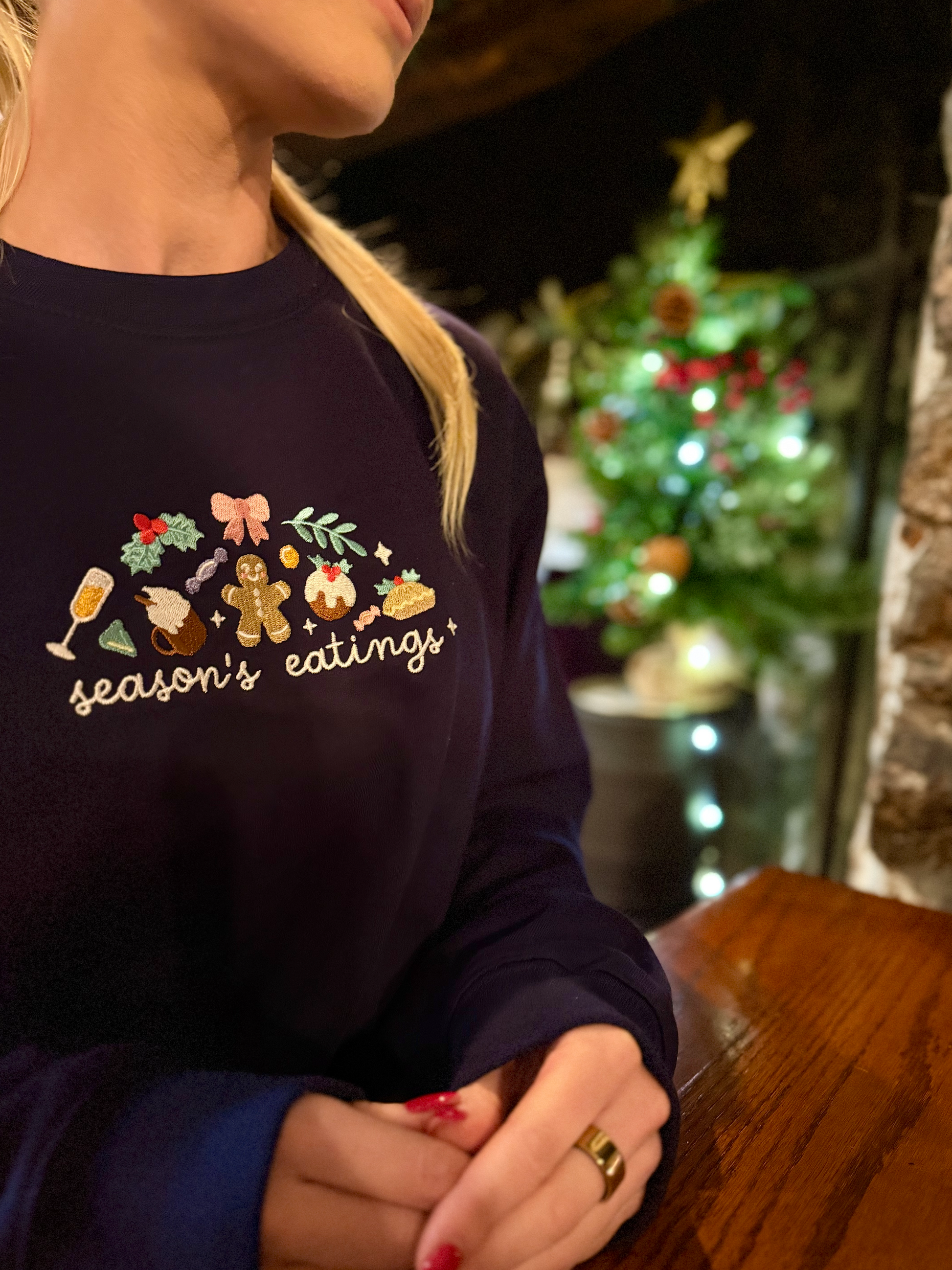 Person wearing a navy blue sweater with 'season's eatings' design in a festive setting.