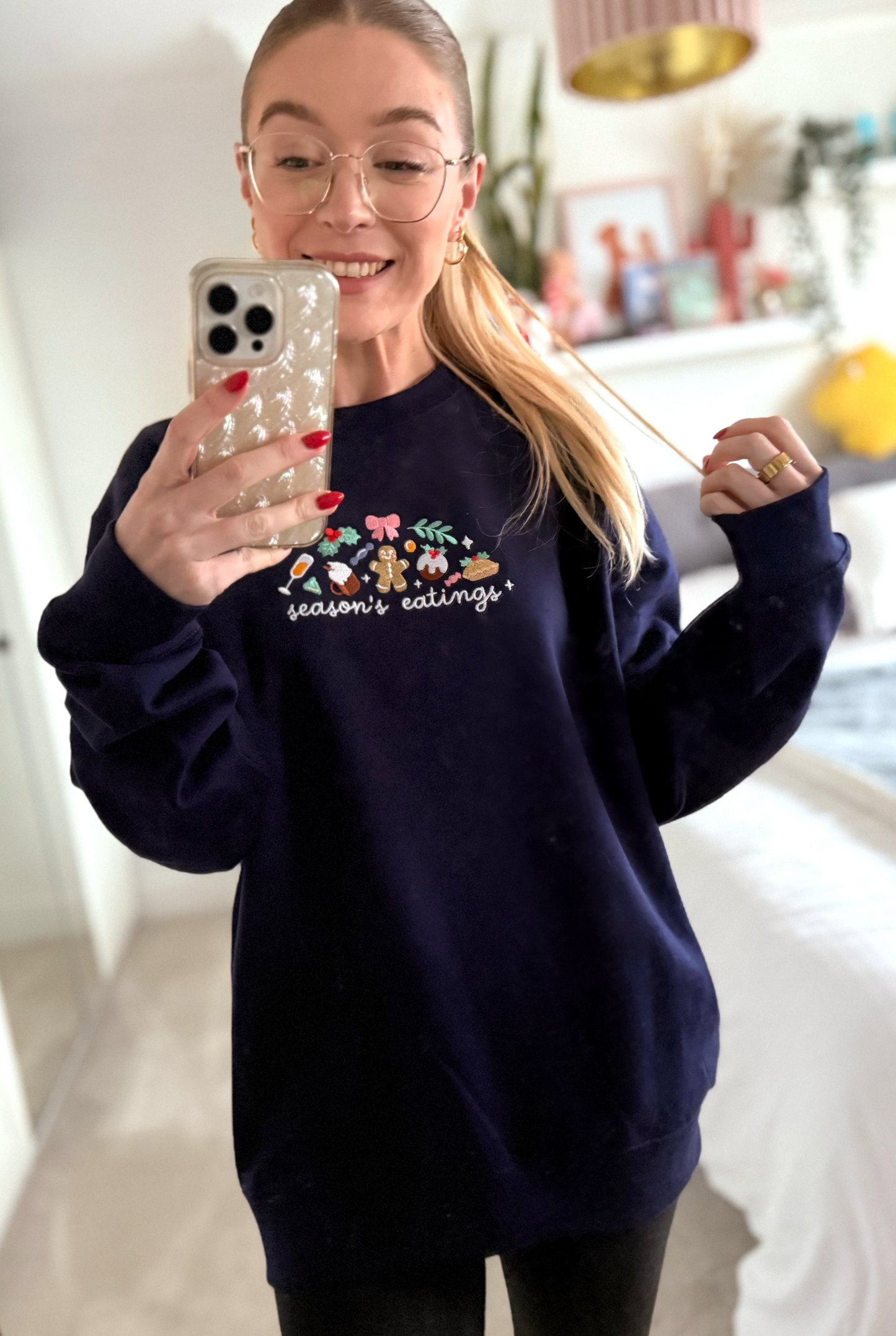 Person wearing a navy sweatshirt with 'season's greetings' text and colorful designs, taking a mirror selfie.
