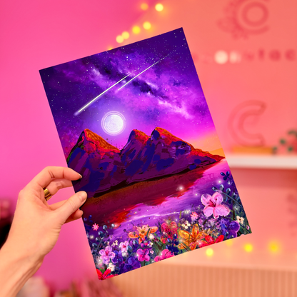 Hand holding a colorful card with a mountain and starry sky design against a pink background.