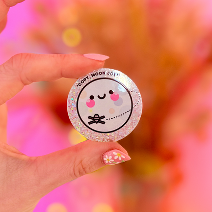Hand holding a round sticker with a moon cartoon character design against a pink blurred background. the text around the moon says "copy, moon joy"