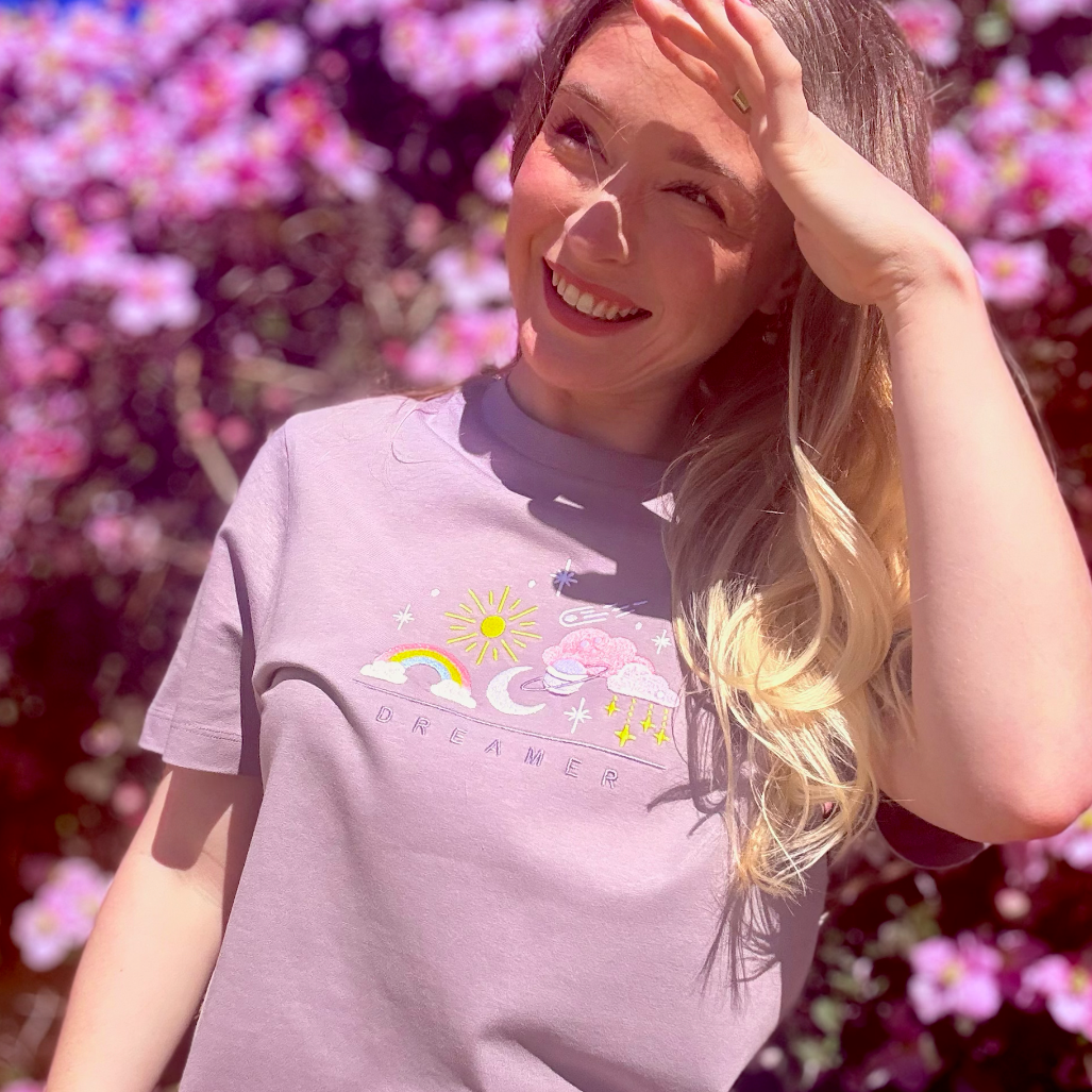 Woman wearing a light purple t-shirt with a colorful graphic in front of pink flowers