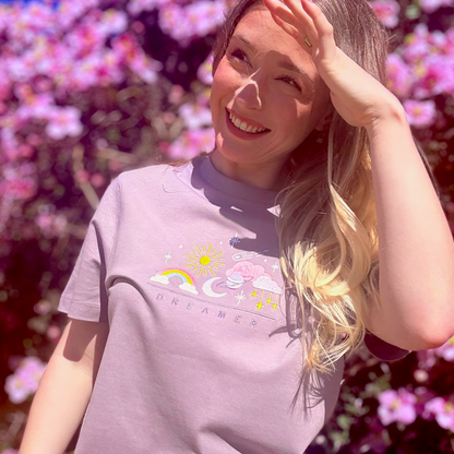 Woman wearing a light purple t-shirt with a colorful graphic in front of pink flowers