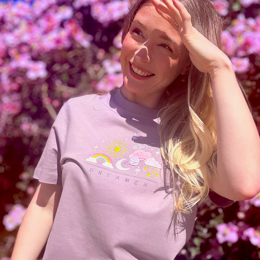 Woman wearing a light purple t-shirt with a colorful graphic in front of pink flowers