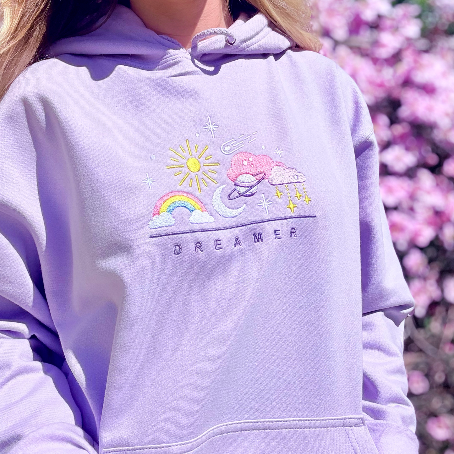 Person wearing a light purple hoodie with a colorful design and 'Dreamer' text, standing against a blurred floral background.