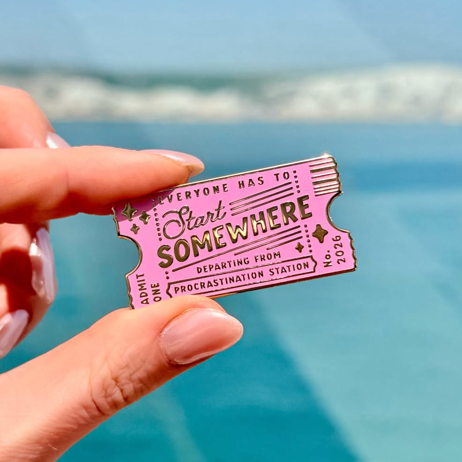 Pink ticket-shaped enamel pin with text saying 'everyone has to start somewhere' held by a hand against a blue sky and water background