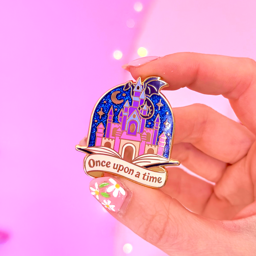 Pin with castle design, with black dragon sat on top, popping out of a book and 'Once upon a time' text held by a hand against a pink background