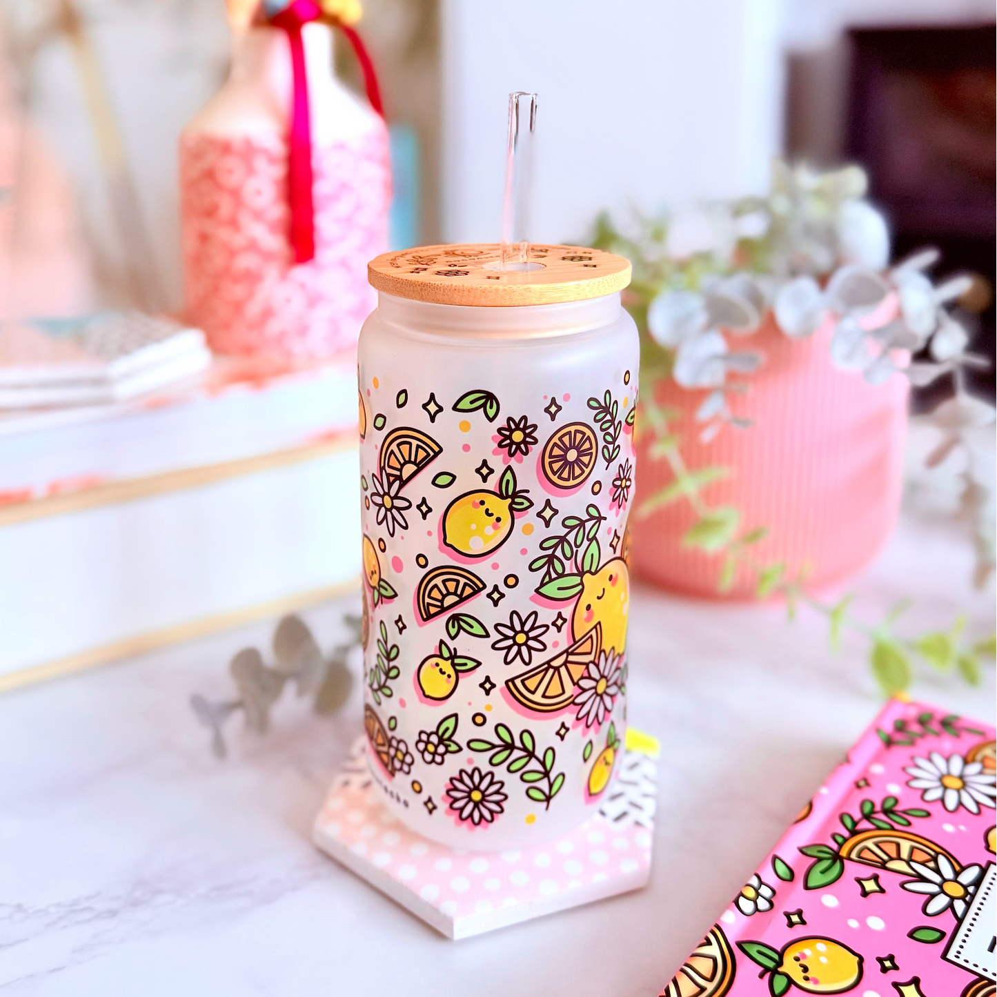 A clear glass can-shaped tumbler with a bamboo lid and metal straw, decorated with a colorful lemon and floral pattern, set on a marble countertop in a styled home environment.