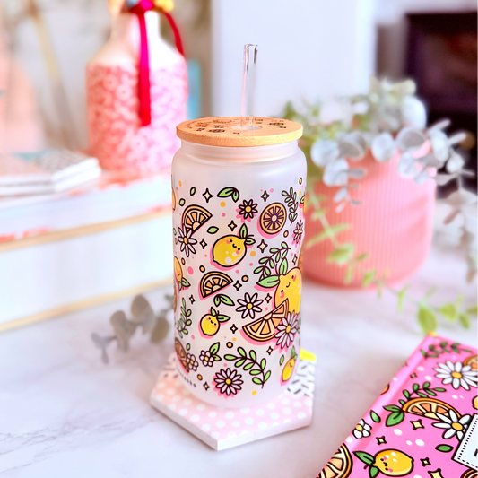 A clear glass can-shaped tumbler with a bamboo lid and metal straw, decorated with a colorful lemon and floral pattern, set on a marble countertop in a styled home environment.