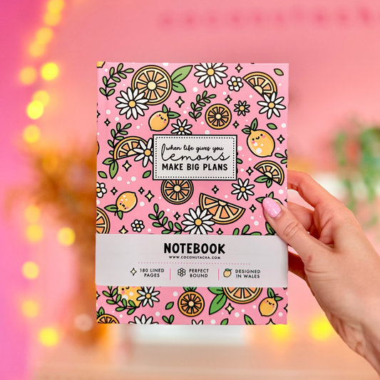 A pink notebook with a lemon and flower pattern is held by a hand against a warm, blurred pink background with bokeh lights.