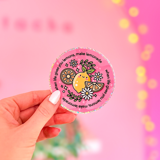 A hand with white manicured nails holds a round pink sticker featuring a cute lemon illustration and the text 'make lemonade, make lemonade, make lemonade' against a bright pink bokeh background.