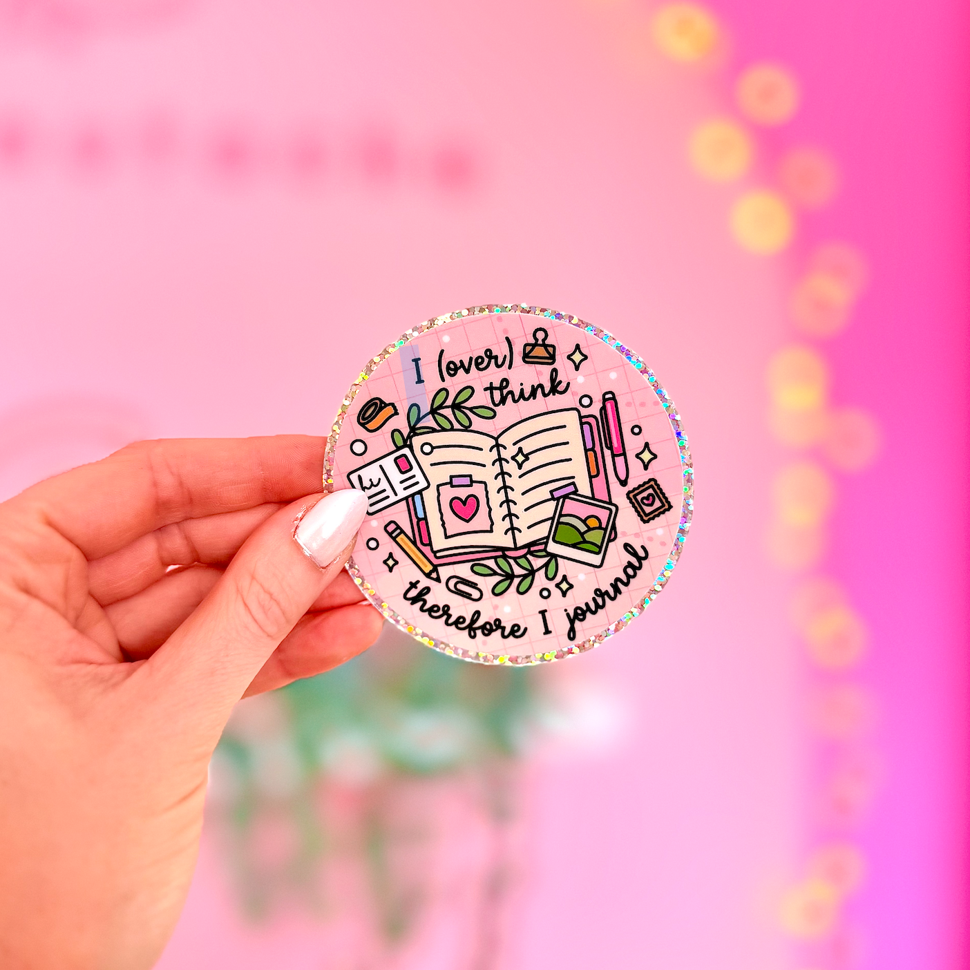 A hand with white manicured nails holds a round sticker featuring a colorful doodle of a notebook, coffee cup, and motivational text that reads 'I love to think therefore I prove'. The background is a bright pink wall with warm bokeh string lights.