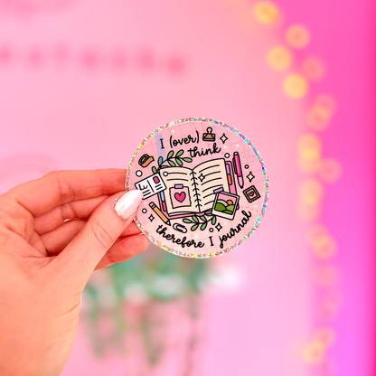 A hand with white manicured nails holds a round sticker featuring a colorful doodle of a notebook, coffee cup, and motivational text that reads 'I love to think therefore I prove'. The background is a bright pink wall with warm bokeh string lights.