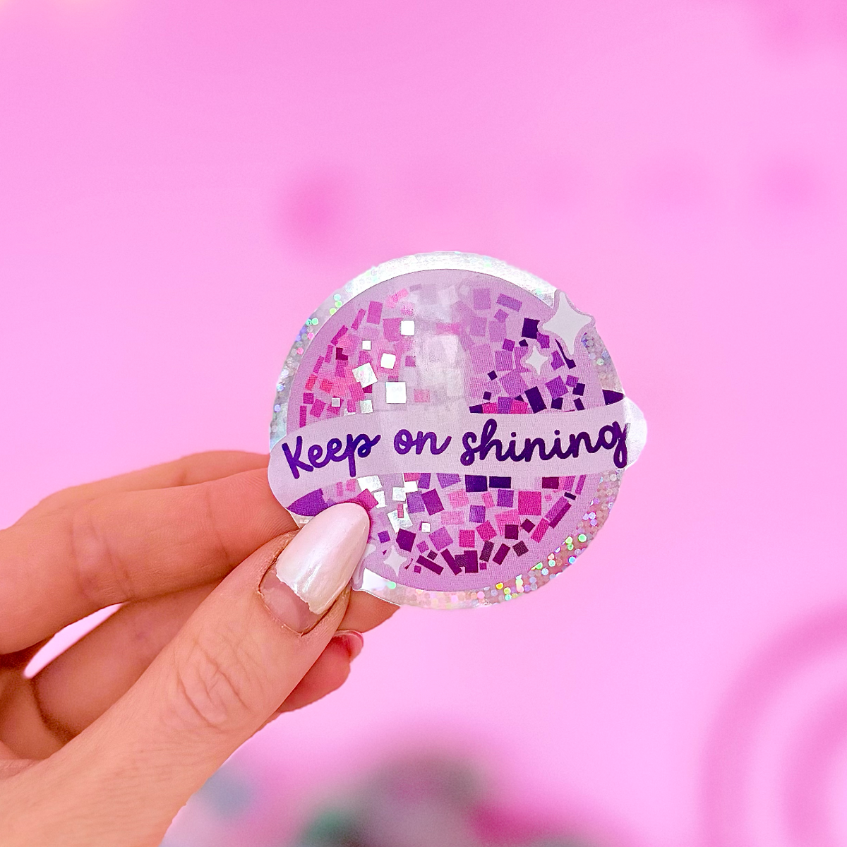 A hand with light pink nail polish holds a round clear sticker featuring a purple peace sign filled with glitter and the phrase 'Keep on shining' in purple script. The background is a bright pink bokeh with soft light spots.