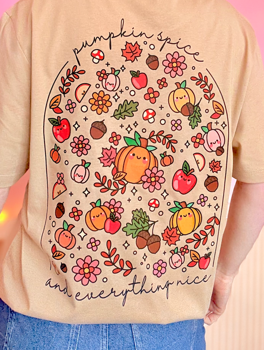 A person wearing a beige short-sleeve T-shirt with a large autumn-themed graphic print on the back. The print features cute illustrations of pumpkins, apples, flowers, leaves, acorns, and mushrooms, with the text 'pumpkin spice' at the top and 'and everything nice' at the bottom.
