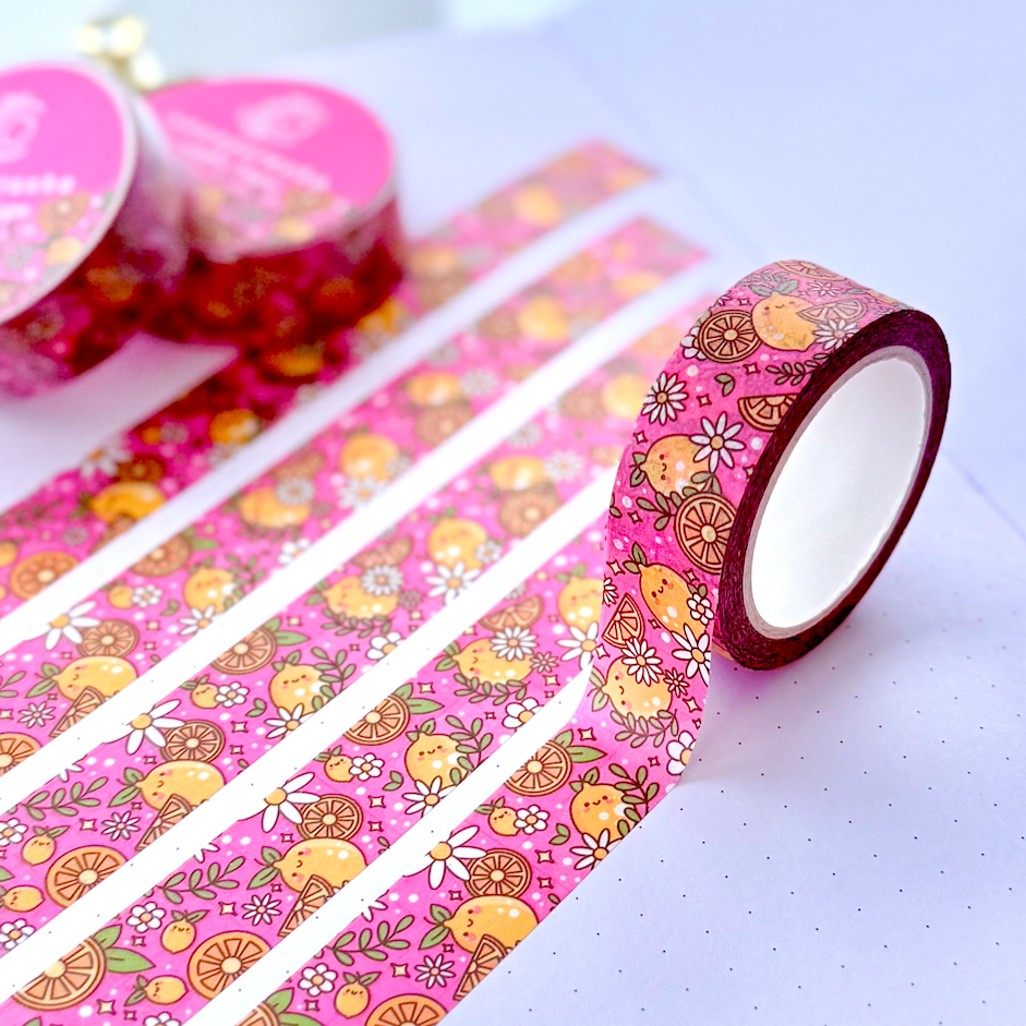 Pink patterned washi tape with cartoon fruit and floral designs, shown partially unrolled across an open notebook with two rolls in the background.