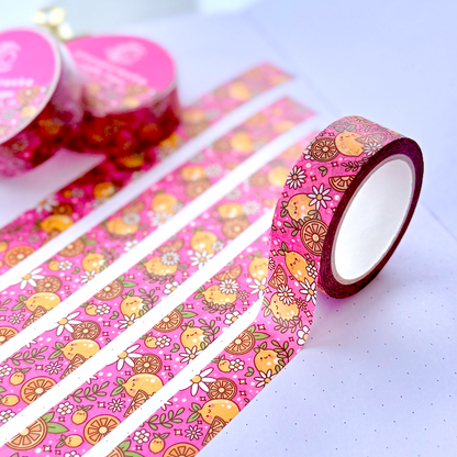 Pink patterned washi tape with cartoon fruit and floral designs, shown partially unrolled across an open notebook with two rolls in the background.