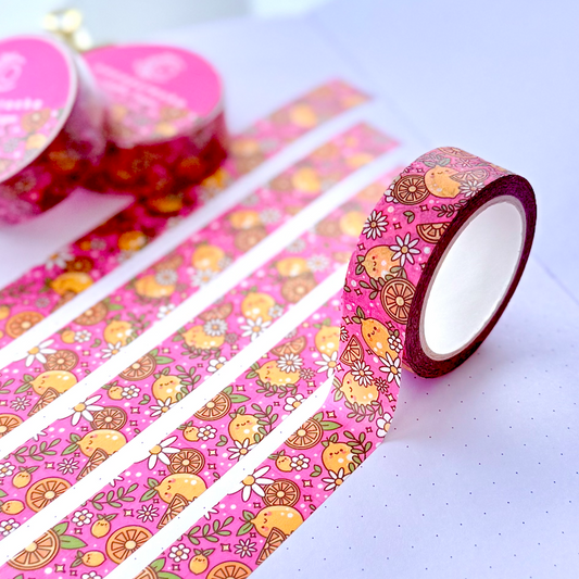 Pink patterned washi tape with cartoon fruit and floral designs, shown partially unrolled across an open notebook with two rolls in the background.