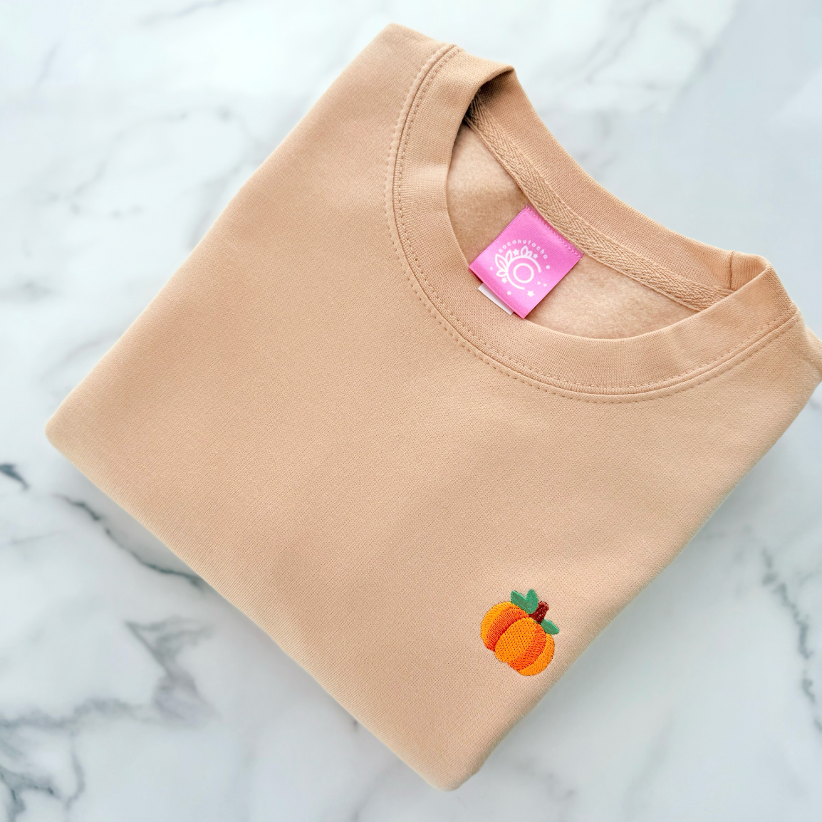 A folded beige crewneck sweatshirt with a small embroidered peach graphic on the lower left chest, displayed on a white and gray marble surface. The inner collar features a pink brand tag.