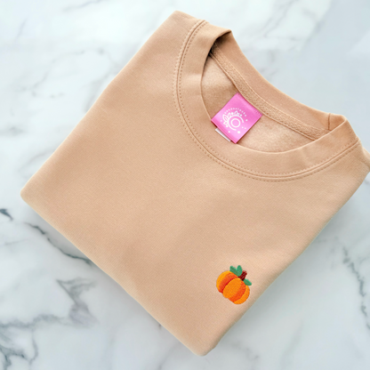 A folded beige crewneck sweatshirt with a small embroidered peach graphic on the lower left chest, displayed on a white and gray marble surface. The inner collar features a pink brand tag.