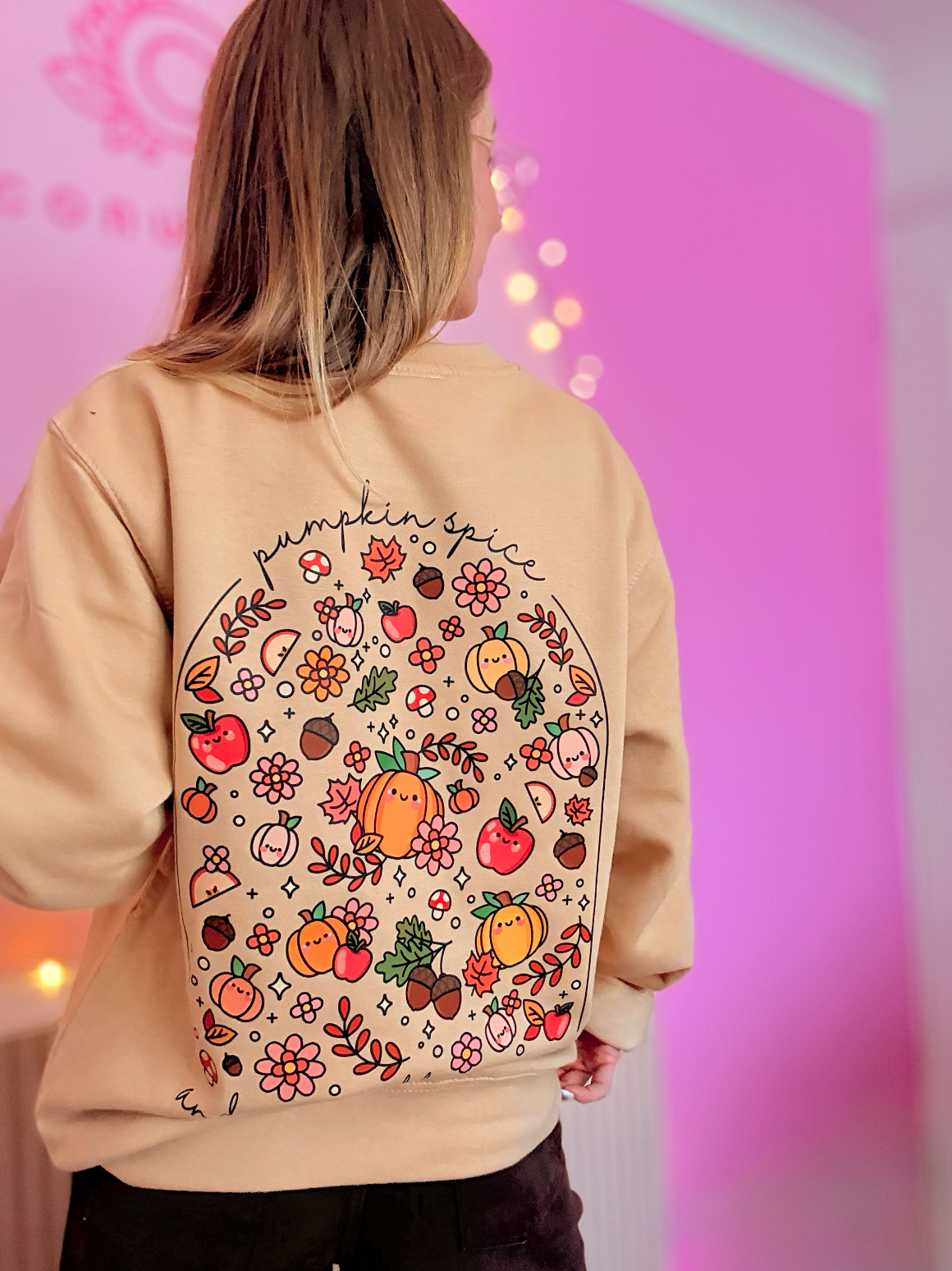 A woman viewed from behind wearing a beige sweatshirt with a large autumn-themed graphic print on the back, featuring pumpkins, acorns, flowers, and the word 'pumpkin pie' in script, against a pink-lit indoor background.