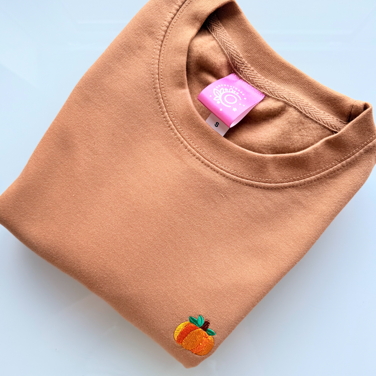 A folded peach-colored crewneck sweatshirt with a small embroidered peach patch on the lower left hem, displayed on a light blue background.