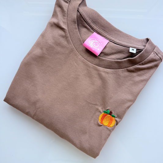 A folded brown crewneck t-shirt with a small embroidered peach patch on the left chest, displayed on a light gray reflective surface.