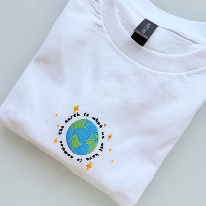 A folded white t-shirt on a light gray reflective surface, featuring a colorful graphic of the Earth surrounded by small yellow stars and the text 'the earth is what we live' in black.