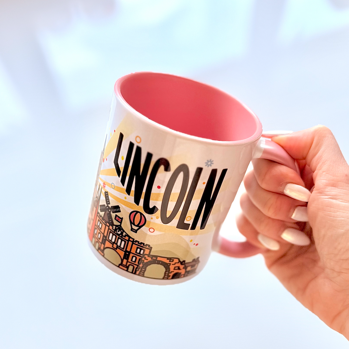 A hand with white manicured nails holds a ceramic mug featuring a pink interior and a cream exterior printed with the word 'LINCOLN' and an illustration of historic Lincoln, Nebraska landmarks.