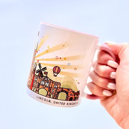A hand with white manicured nails holds a white ceramic mug featuring a colorful illustration of Lincoln, United Kingdom, against a soft light blue background.