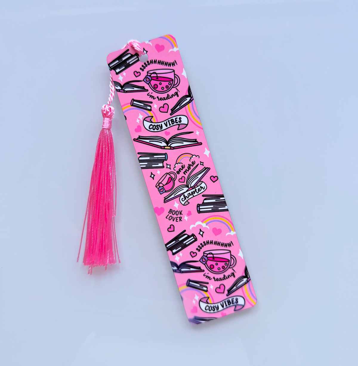 A pink fabric bookmark with a matching tassel, decorated with black and white illustrations of books, coffee cups, rainbows, and text like 'COFFEE VIBE' and 'BOOK & LOVER', shown on a light blue background.