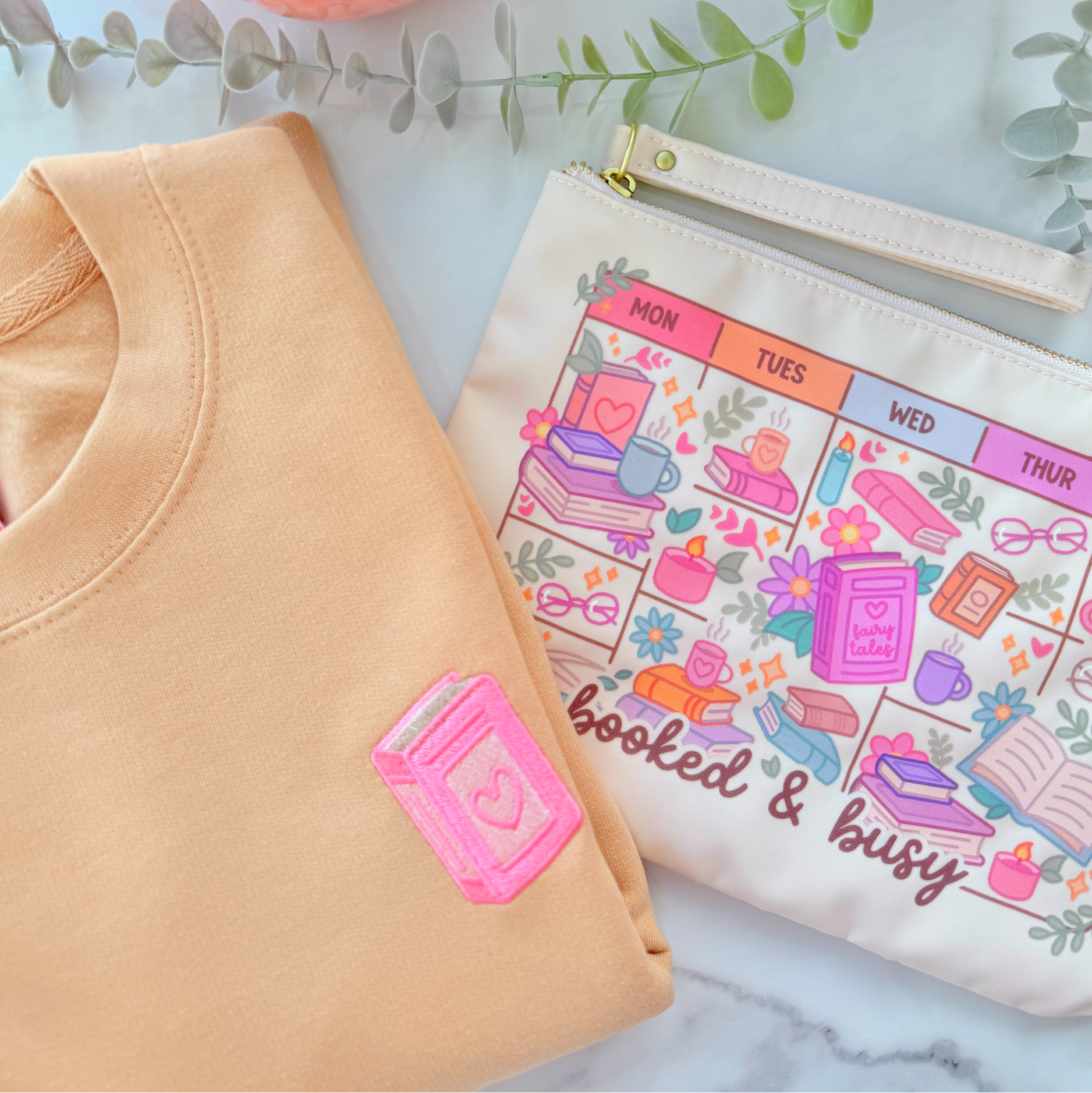 Beige-colored pouch with gold zipper and 'booked & busy' design on a marble surface with a pink mug and greenery.