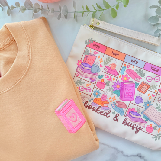 Beige-colored pouch with gold zipper and 'booked & busy' design on a marble surface with a pink mug and greenery.