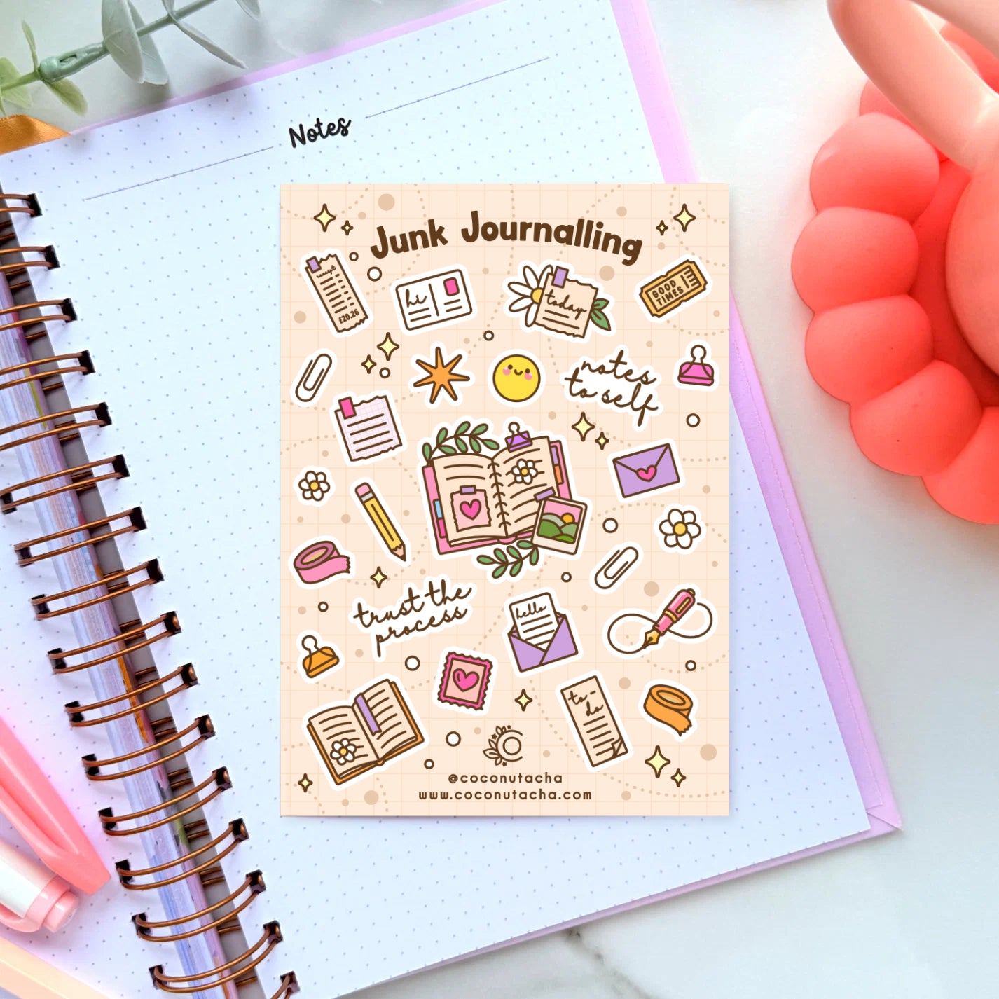 A rectangular planner sticker sheet titled 'Junk Journalling' with cute doodle illustrations and phrases like 'notes to self' and 'trust the process,' placed on an open lined notebook. The scene is styled with a coral phone, green leaves, and a pink pen.