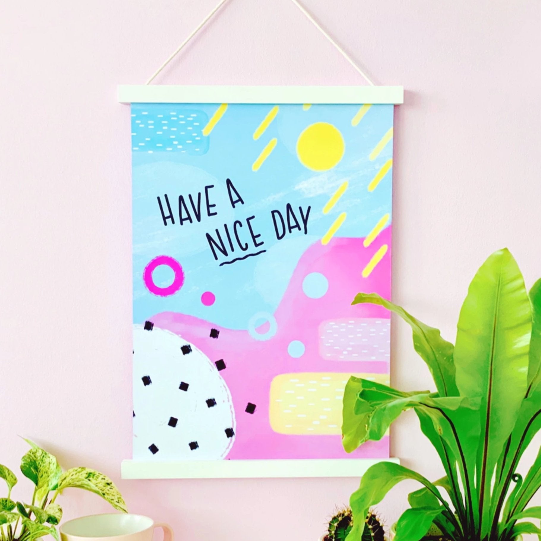 A brightly coloured art print placed on a pastel pink wall, surrounded by lush green plants in brightly coloured plant pots. The art print consists of abstract shapes and brightly coloured blues, pinks and yellows. There's a quote in a handwritten font saying 'have a nice day'.